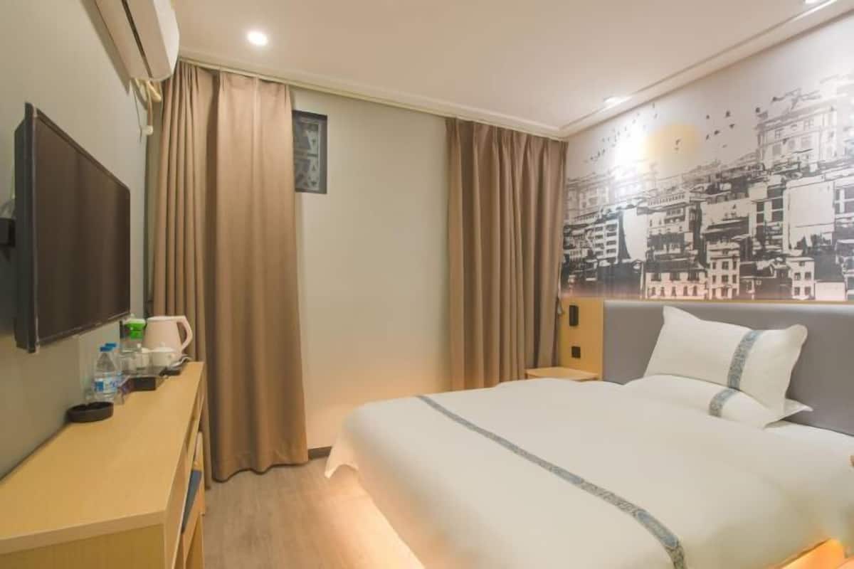 A hotel room with a bed, a desk with a coffee maker, curtains, a wall-mounted TV, and a cityscape mural.