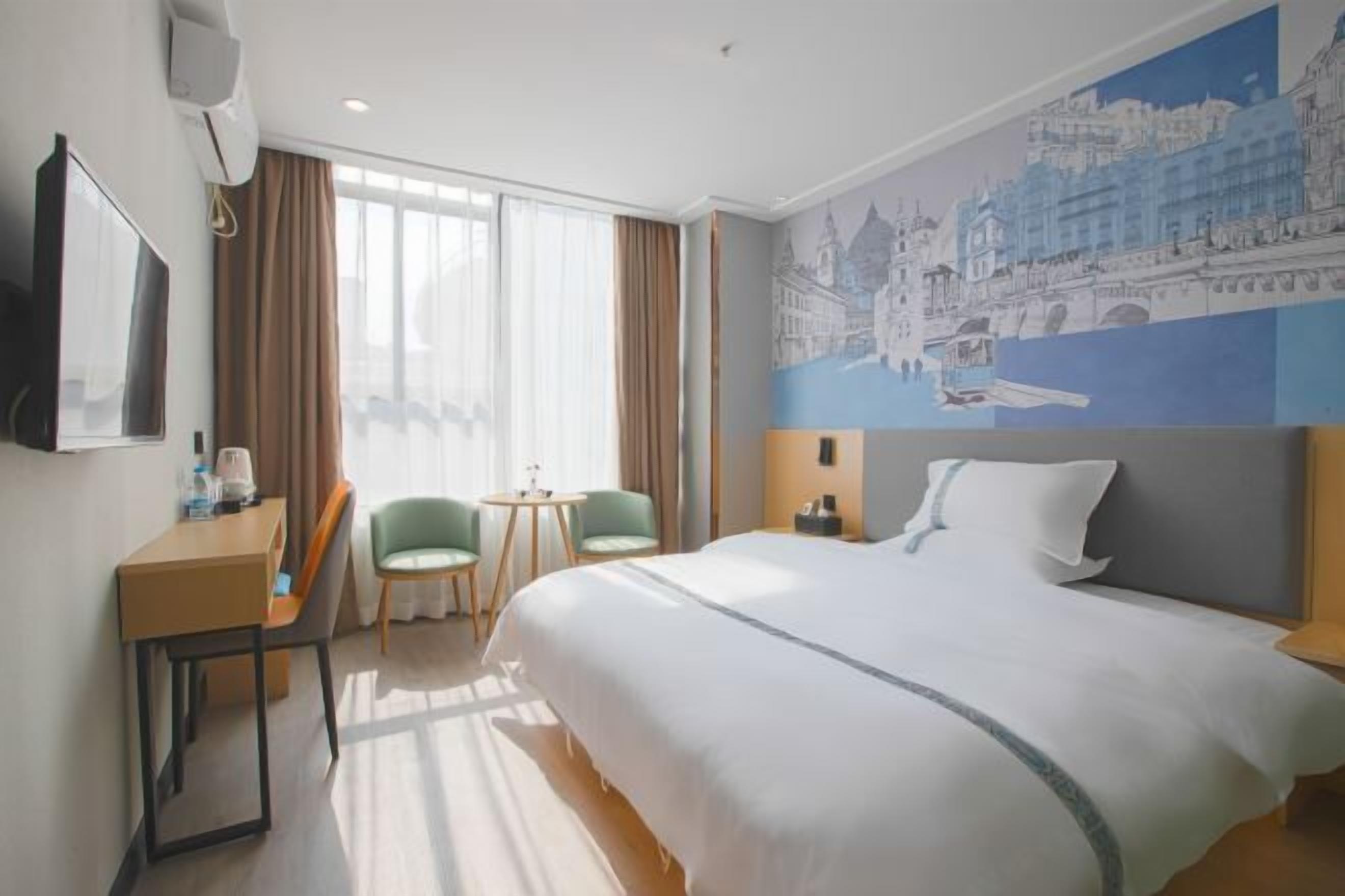 Quarto de hotel com uma cama grande, uma escrivaninha com cadeira, uma mesinha com duas cadeiras e uma janela ampla com cortinas.