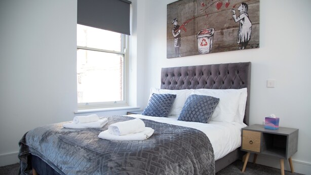 A bedroom with a bed, a window, a nightstand, and a painting on the wall. - Hotel Rafay (Blackburn)