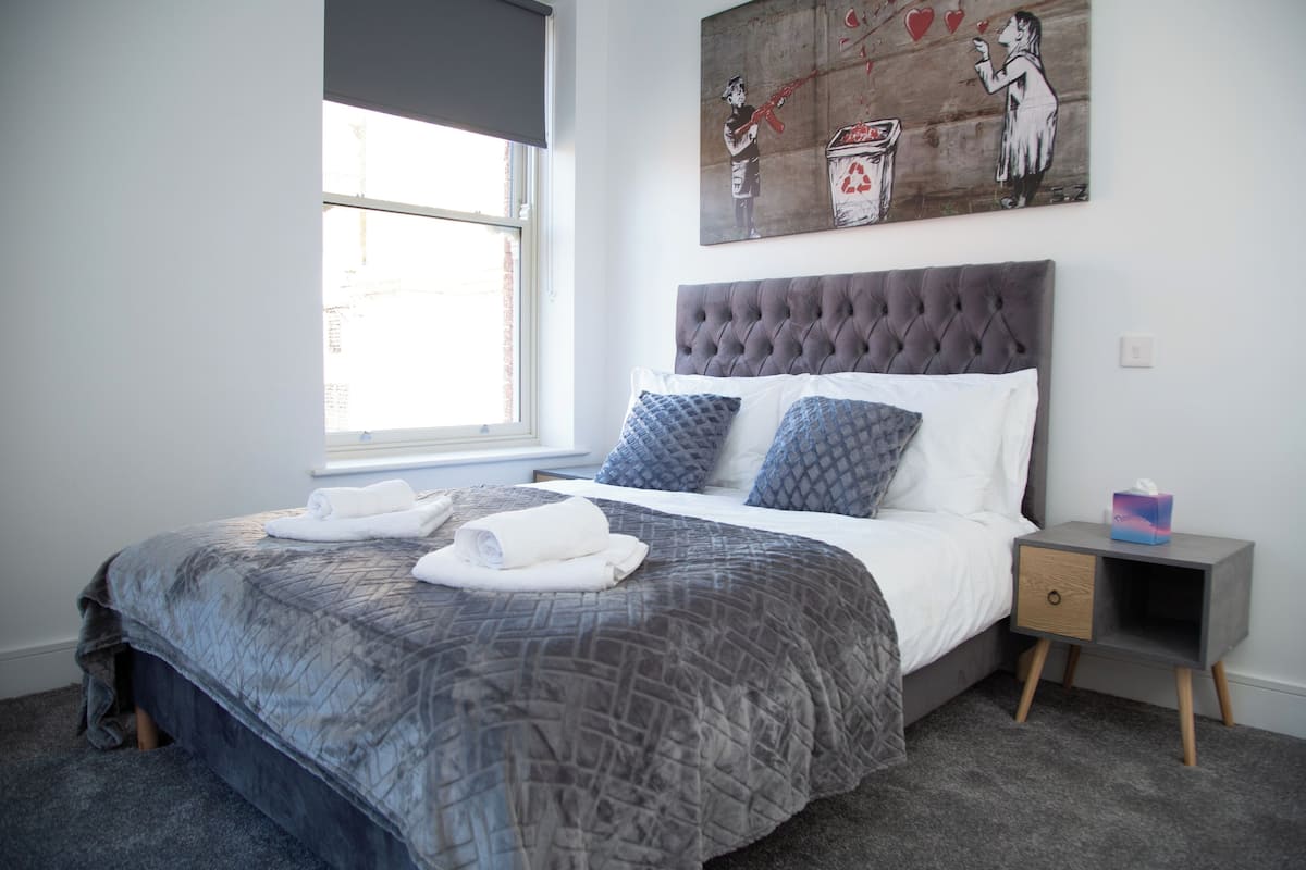 Un dormitorio con cama, ventana, mesita de noche y un cuadro en la pared.