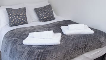 Una cama bien hecha con ropa de cama gris y dos toallas blancas, una pared con un cuadro de mariposas coloridas y almohadas con estampado gris.