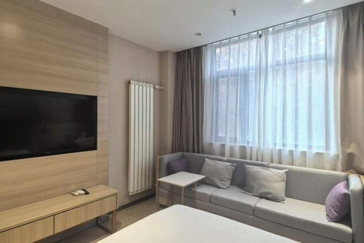 Ein modernes Hotelzimmer mit Flachbildfernseher, einer Couch, einem kleinen Tisch und einem Fenster mit transparenten Vorhängen.