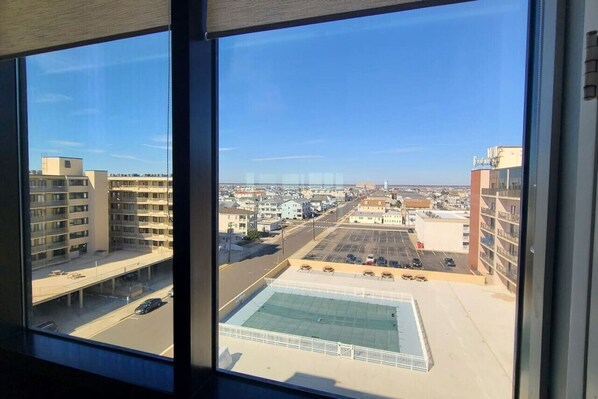 A view from a high-rise window overlooking a cityscape with multiple buildings, a parking lot, and a swimming pool. - Ocean-Front Penthouse 3BR/2BA/7Bed @ Regency Tower (North Wildwood)