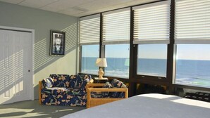 Una habitación con cama, sofá floral, lámpara y ventanas amplias con vistas al océano.