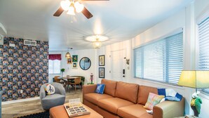A cozy living room with a brown leather sofa, a grey armchair, and a black rug. There's a ceiling fan, a chandelier, and a window with blinds. - The Hutt, 2BR Chic Condo by Velvet Ditch Villas (Oxford)