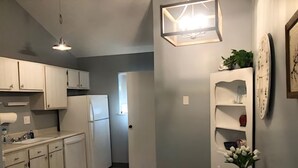 A compact kitchen with white cabinets, a refrigerator, a wooden table, and a wall-mounted shelf with decorative items.