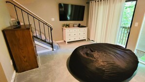 A room with a staircase, a flat-screen TV, a white dresser, a bean bag chair, and a balcony with curtains.