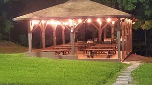 A covered pavilion with picnic tables and barbecue grills, surrounded by grass and trees.