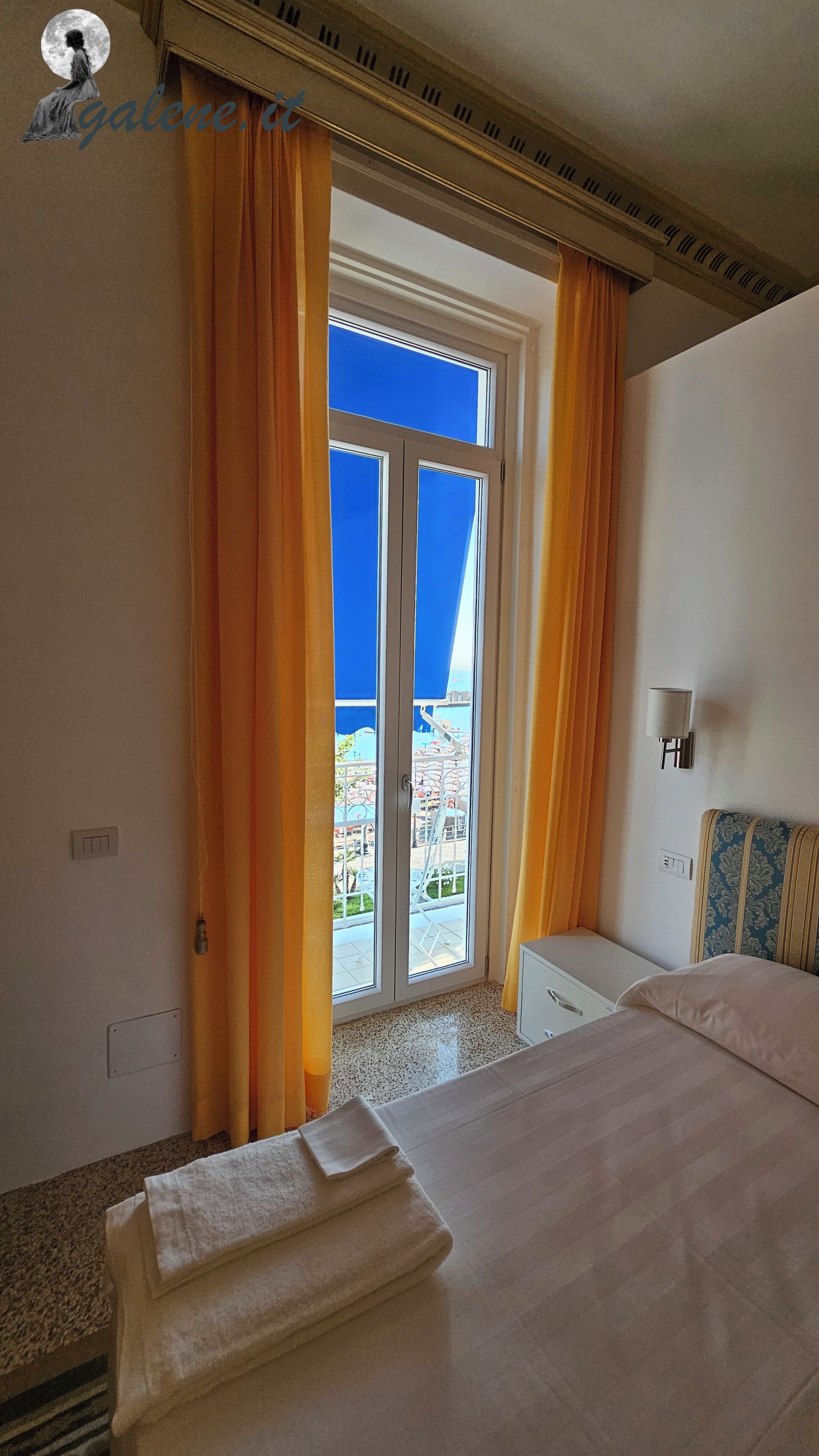 A hotel room with a bed, a desk, and a view of a building through a glass door.