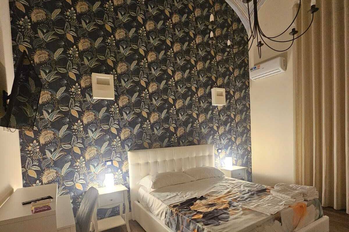 A bedroom with a large bed, a desk, and a chair. There is a patterned wallpaper on the wall and a chandelier hanging from the ceiling.