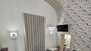 Habitación de hotel con zona de comedor, televisor montado en la pared y papel tapiz estampado.