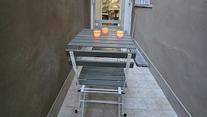 Un estrecho pasillo exterior con una mesa de metal y bancos, una ventana con barrotes blancos y una puerta que da a un espacio interior.