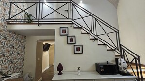 Una escalera con barandilla de metal negro, una pared blanca con cuadros enmarcados y una zona de cocina con microondas y cafetera.