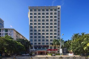 A multi-story hotel with a uniform facade, located at a street corner with palm trees and other buildings in the background. - Xinxing Garden Hotel (Sanyawan) (Sanya)