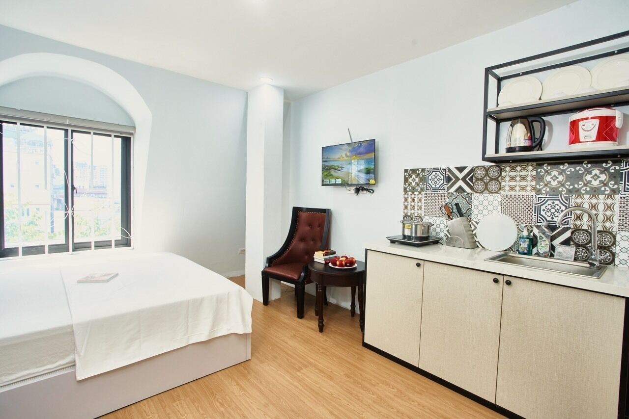 A compact room with a bed, a dining table, a chair, a kitchen area with a sink, and a TV mounted on the wall.
