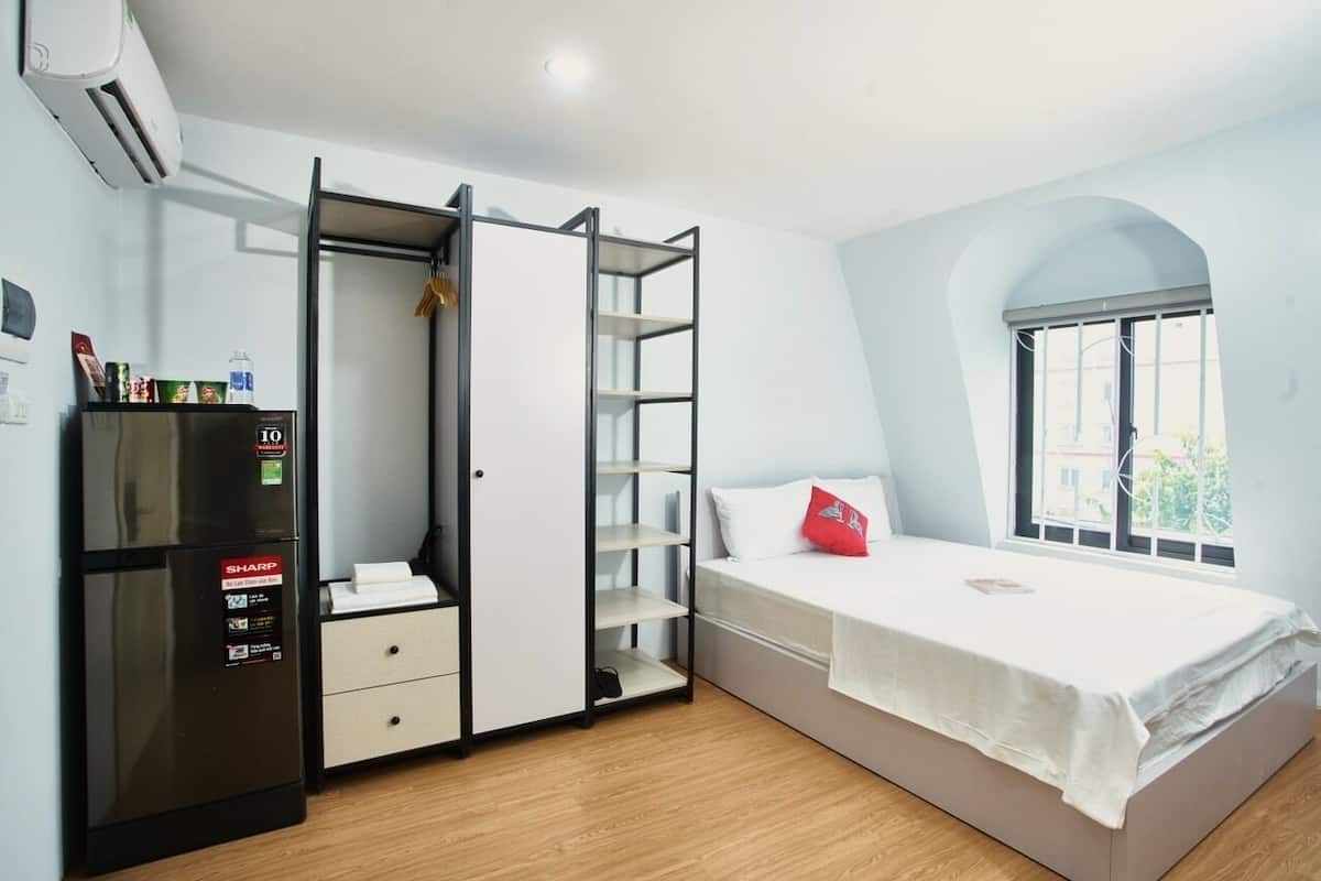 A compact bedroom with a bed, a wardrobe with shelves, a small desk, and a window.