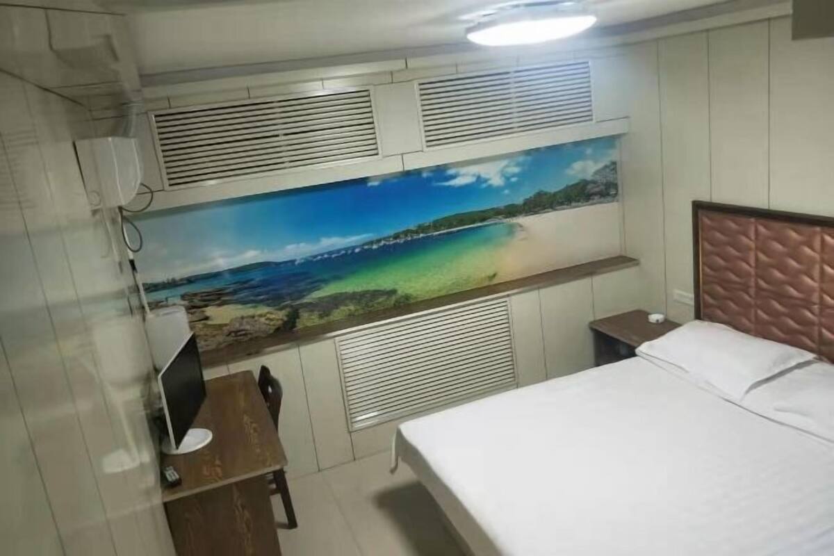 A bedroom with a bed, a desk with a computer, a wall-mounted air conditioner, and a large beach mural.