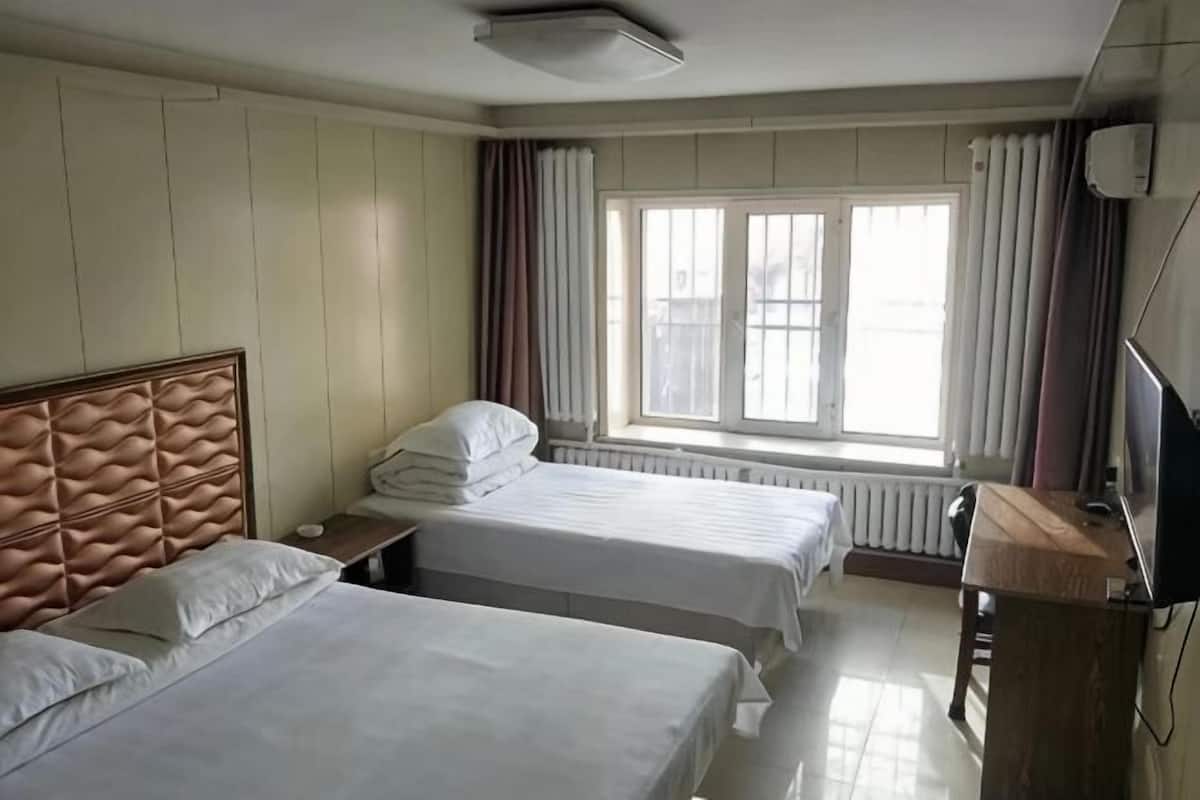 A hotel room with two beds, a wooden headboard, a window with curtains, a radiator, and a desk with a chair.