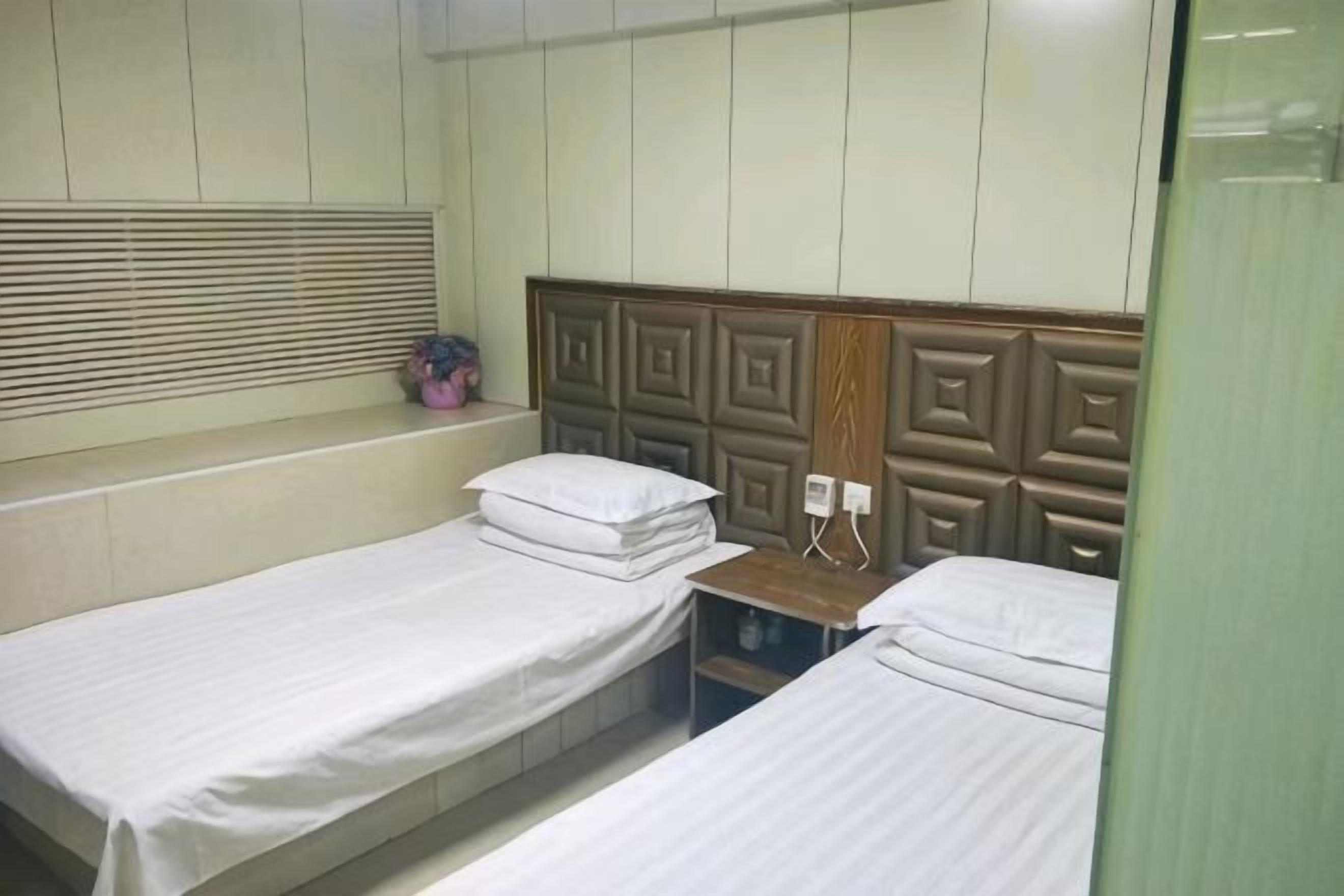 A room with two single beds, a wooden headboard, a window with blinds, and a small shelf with a plant.