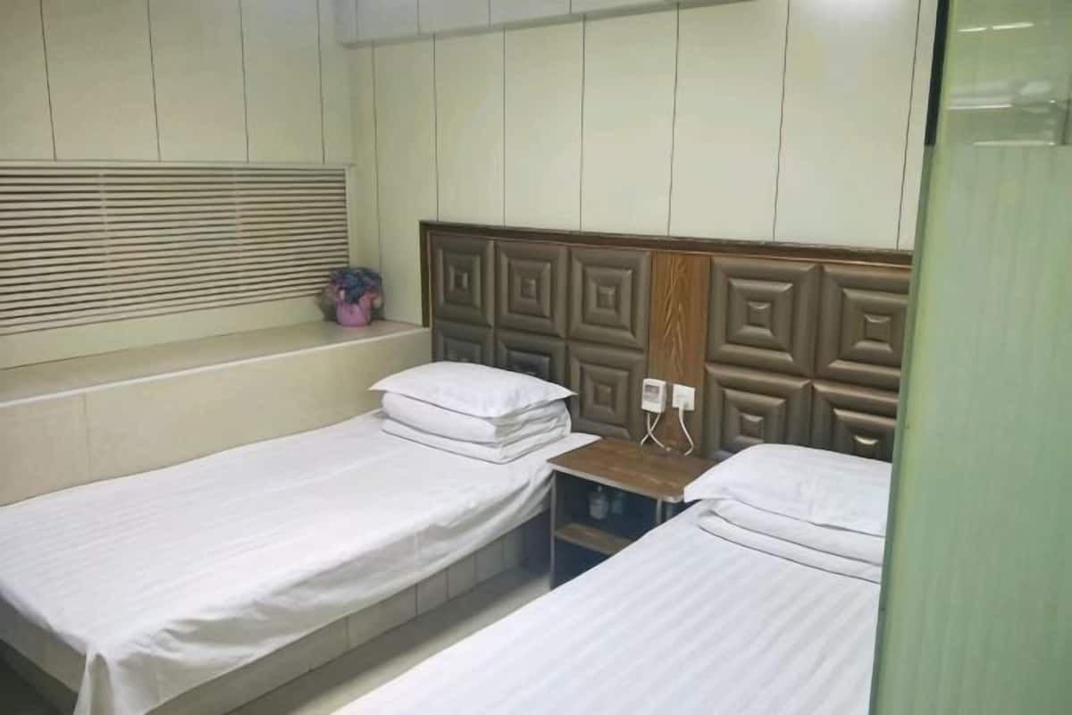 A room with two single beds, a wooden headboard, a window with blinds, and a small shelf with a plant.
