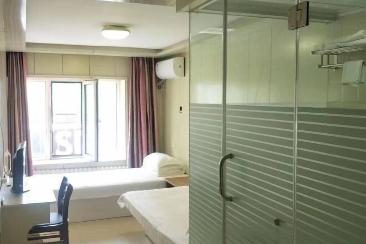 A hotel room with a bed, a desk, a chair, a window with curtains, and a glass partition.