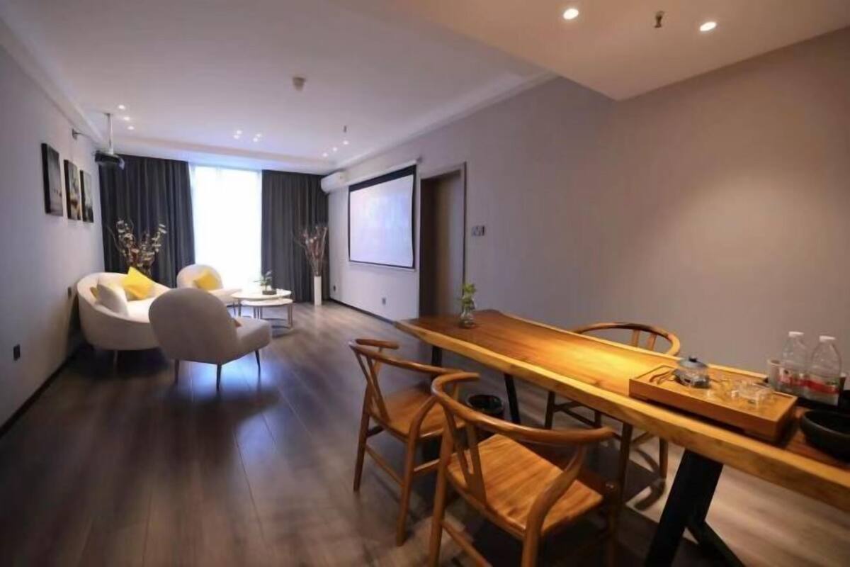 A modern hotel room with a wooden dining table, two chairs, a sofa with yellow cushions, a small side table, and a projector screen on the wall.