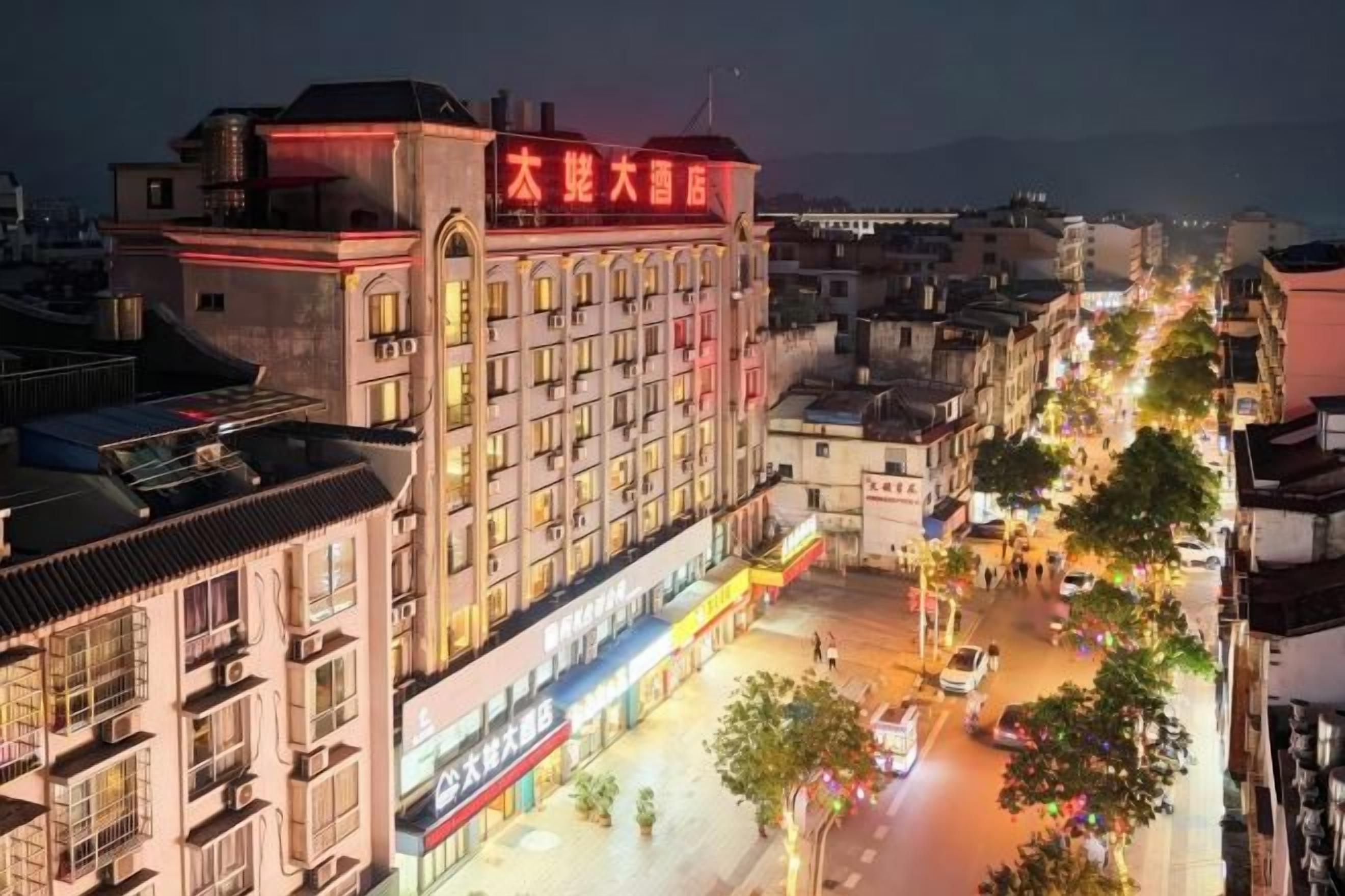 A multi-story hotel with a prominent sign, a street in front with trees and shops, and a clear view of the sky.