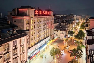 Un hotel de varios pisos con un letrero llamativo, una calle frente al edificio con árboles y comercios, y un cielo despejado.