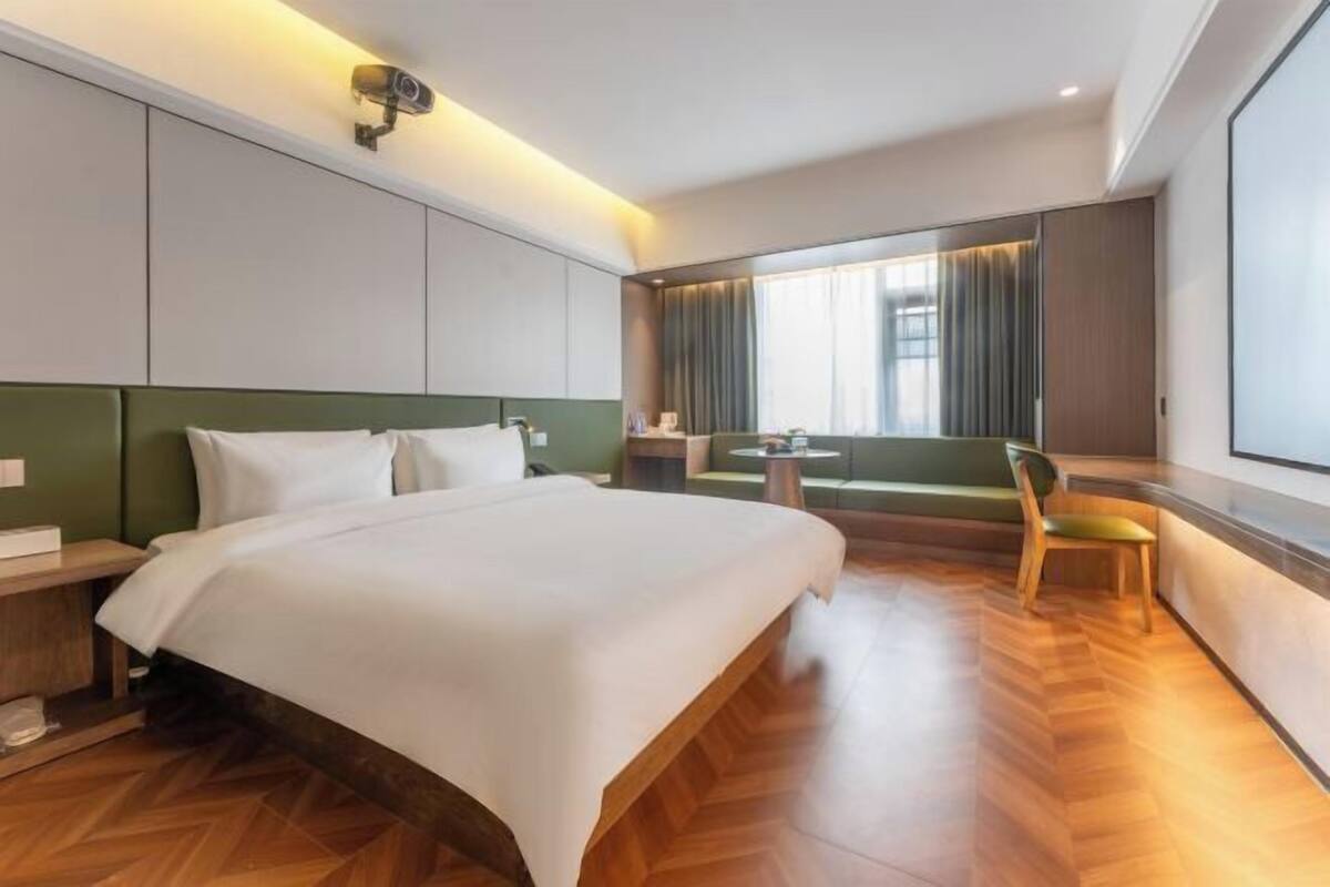 A hotel room with a bed, a small table, a chair, a window with curtains, and a security camera.