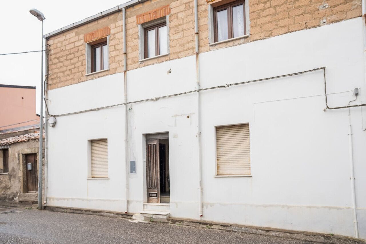 Ein zweistöckiges Gebäude mit weißer Fassade und einem oberen Bereich aus Ziegelsteinen. Im Erdgeschoss befinden sich zwei Fenster und eine Tür mit einer kleinen Stufe zum Eingang.