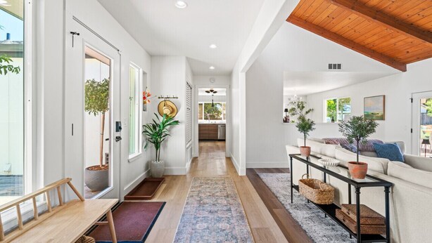 Un intérieur moderne avec un couloir menant à une cuisine, comprenant un banc en bois, des plantes en pot et un canapé avec des coussins.