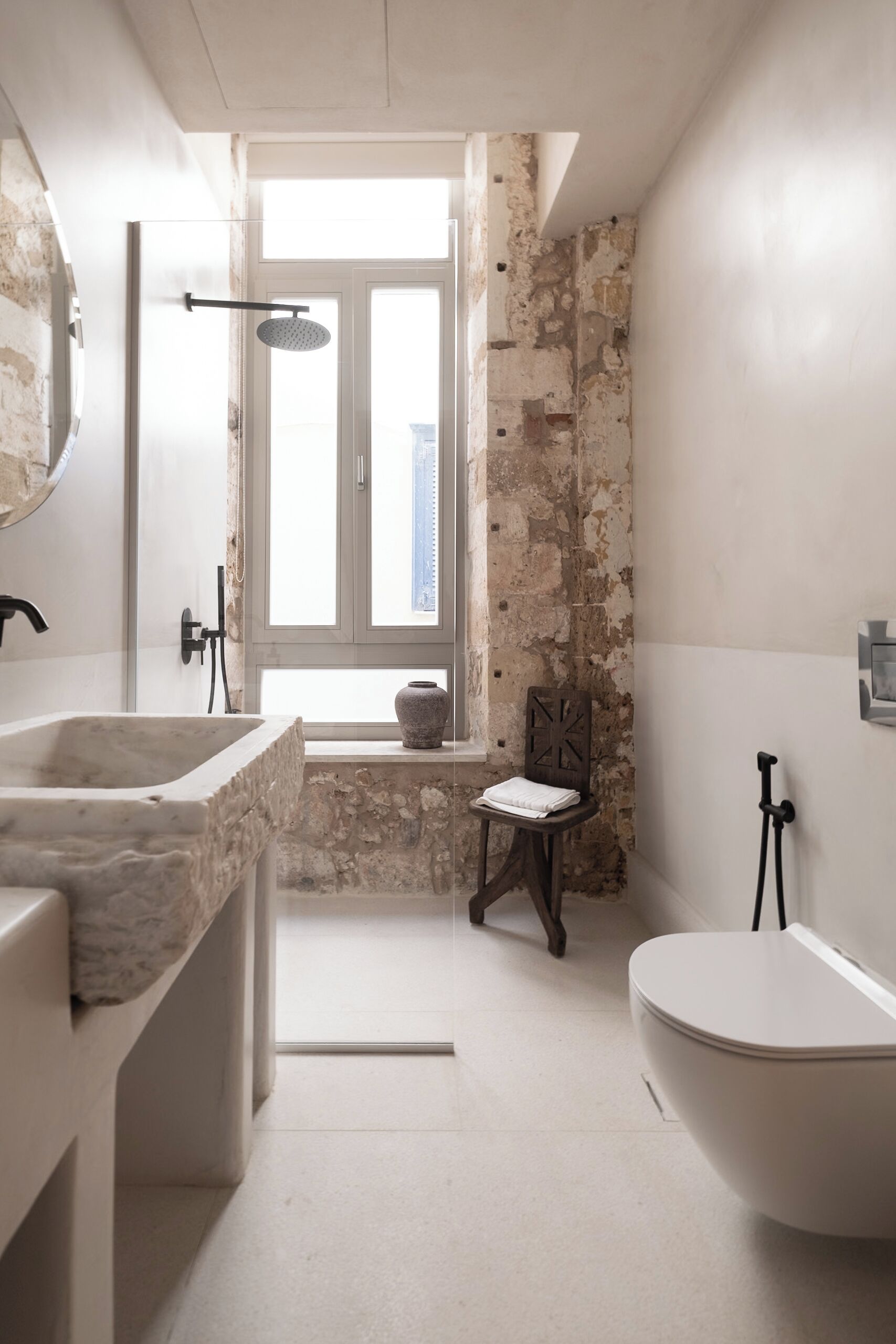 A bathroom with a stone wall, a window, a sink, a toilet, and a chair.