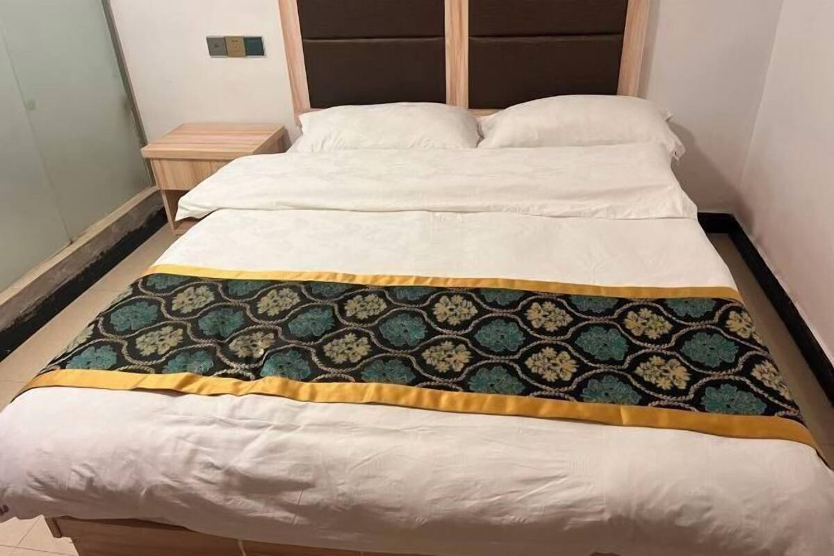 Ein Bett mit gemustertem Überwurf und Kissen, ein Nachttisch aus Holz und eine wandmontierte Steuerung.
