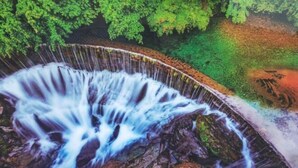 Ein Wasserfall stürzt über Felsen, mit einer geschwungenen Struktur und dichtem Grün im Hintergrund.