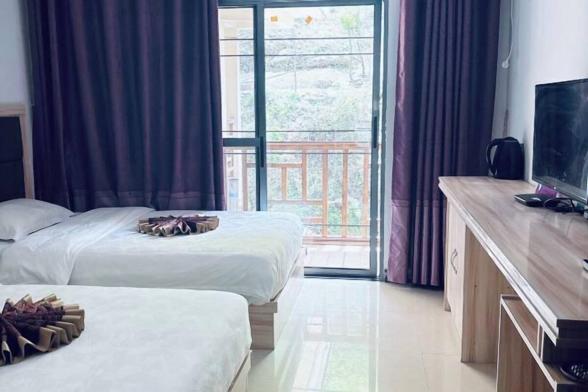 Ein Hotelzimmer mit zwei Betten, einem Fernseher, einem Schreibtisch und einem Balkon mit Vorhängen.