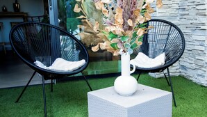 Ein moderner Außenbereich mit zwei schwarzen Stühlen, einer weißen Vase mit Trockenpflanzen und einem strukturierten Tisch auf Kunstrasen.