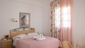 A neatly made bed with a pink bedspread, a framed picture on the wall, a bedside lamp, and a floral curtain. - Marias Hospitality.Pure Paroshospitality (ΠΑΡΟΣ)