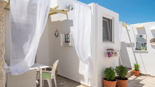 A small outdoor seating area with a table and chairs, a window with shutters, and potted plants. - Marias Hospitality.Pure Paroshospitality (ΠΑΡΟΣ)