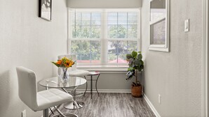 Ein Eckzimmer mit Glastisch, zwei weißen Stühlen, einer Blumenvase, einem Fenster mit Jalousien und einer Pflanze.