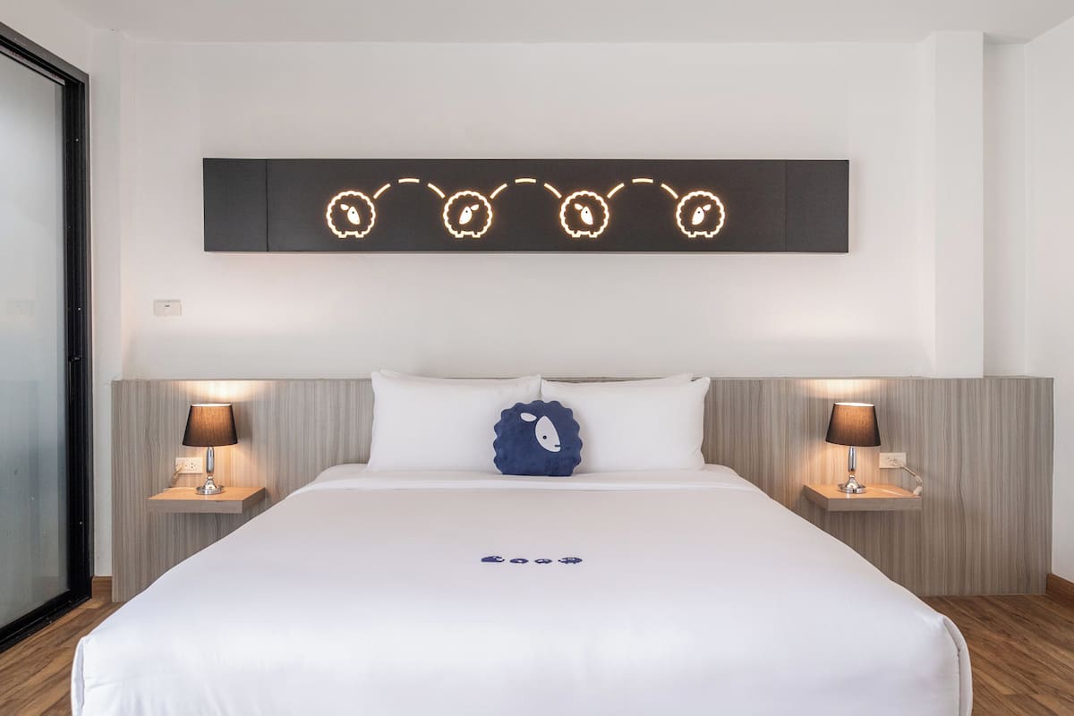 Habitación de hotel con una cama grande, dos mesitas de noche con lámparas, una lámpara de pared con siluetas de animales y una almohada azul con diseño de oveja.