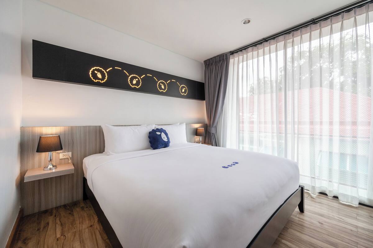 Una habitación de hotel con una cama grande, una mesita de noche con lámpara, una lámpara de pared y cortinas traslúcidas.