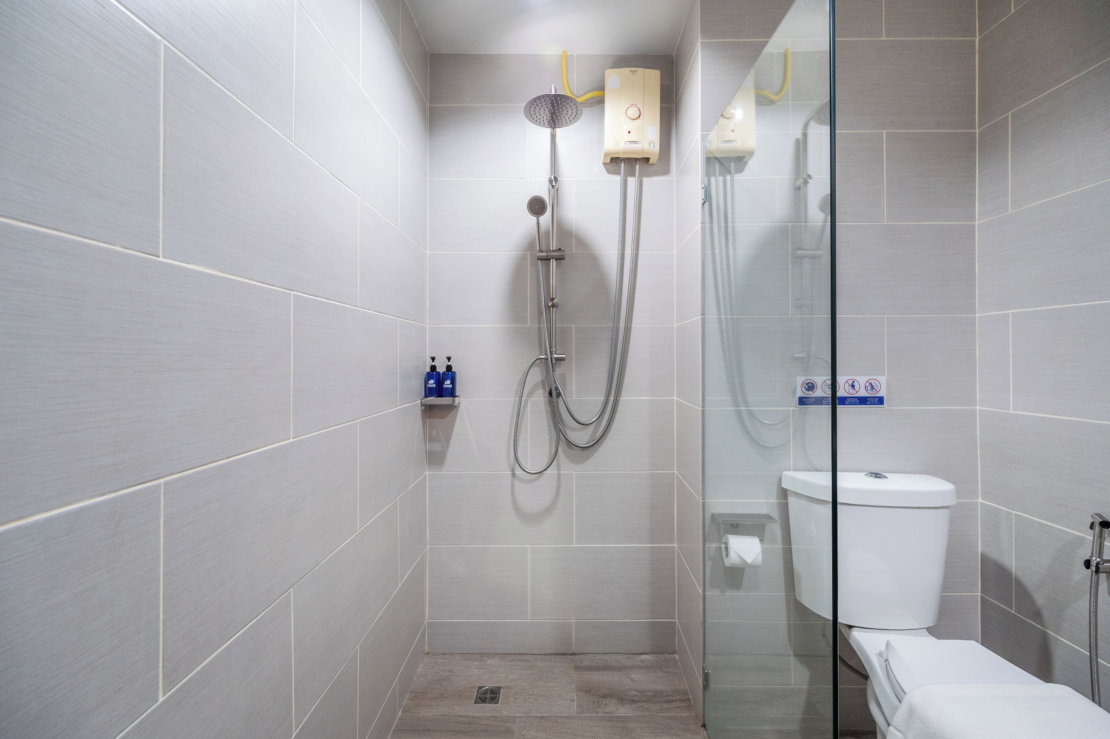 A modern bathroom with grey tiles, a glass shower enclosure, a white toilet, and a small shelf with toiletries.