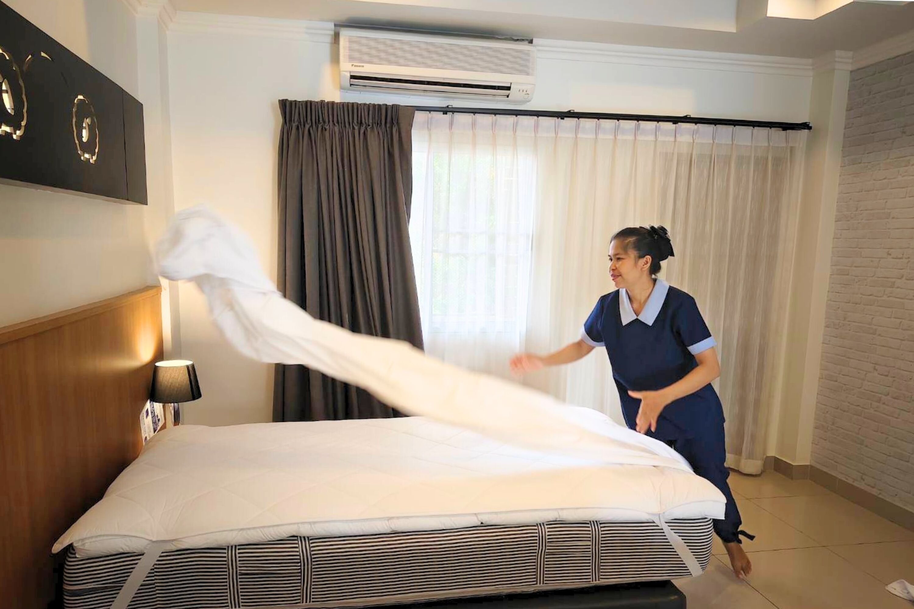A hotel room with a neatly made bed, a wall-mounted air conditioner, and a window with curtains.