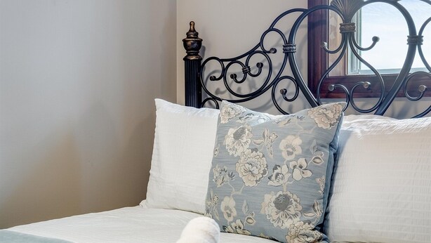 A neatly made bed with white and blue linens, a rolled white towel, and a decorative pillow with a floral pattern.