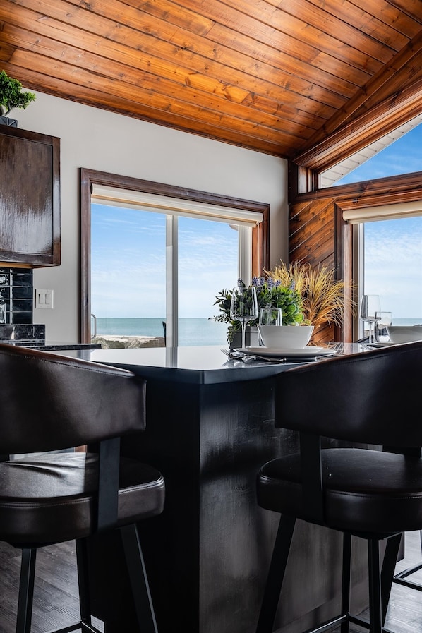 Cucina moderna con soffitto in legno, ampie finestre con vista sul mare e un angolo bar con due sgabelli.