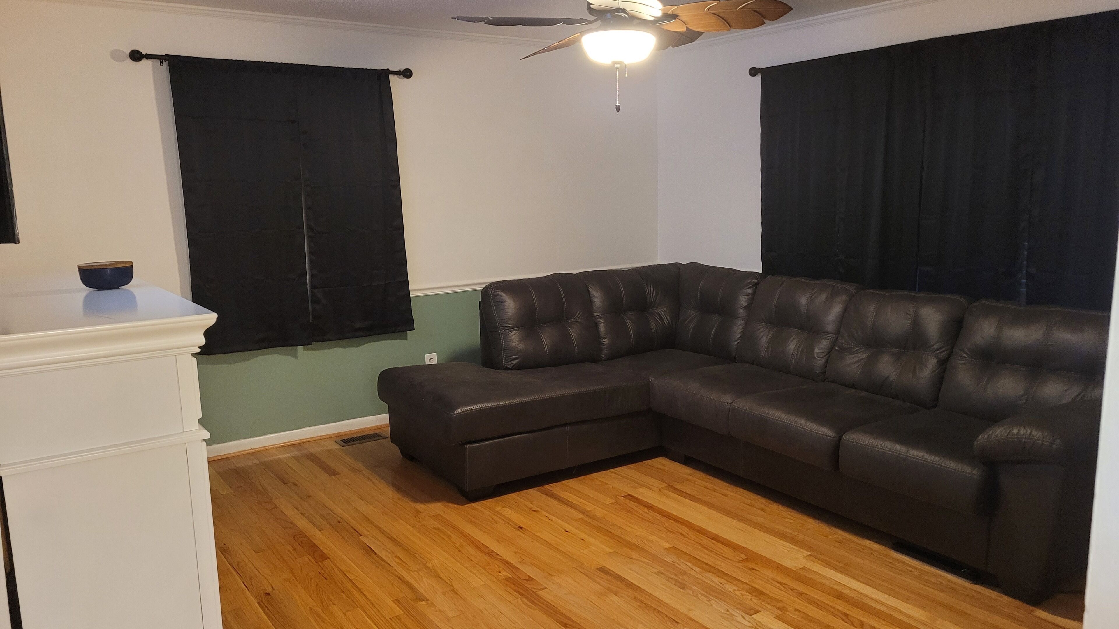 A living room with a sectional sofa, a ceiling fan, and black curtains.