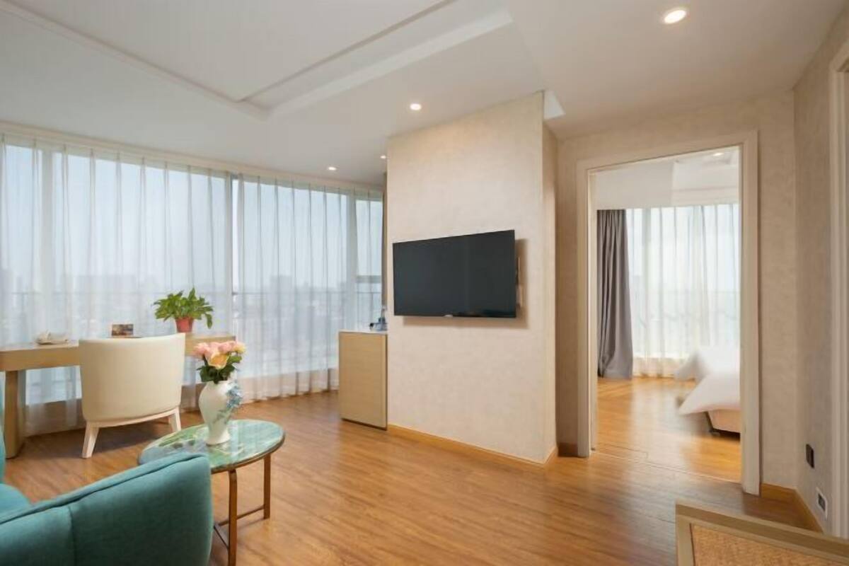 A modern hotel room with a flat-screen TV mounted on the wall, a dining area with a table and chairs, and a cozy seating area with a sofa and a small table with a vase of flowers.