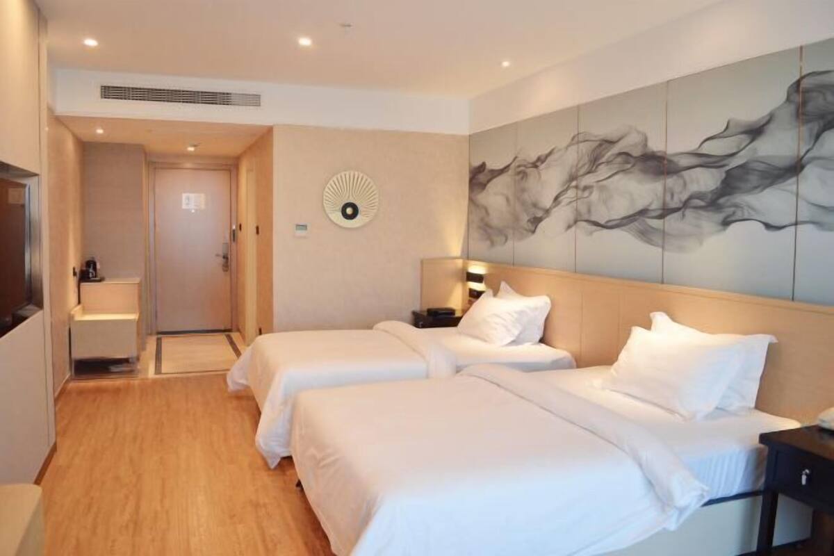 A hotel room with two beds, a large abstract painting on the wall, a bedside table with a lamp, and a view of the hallway.