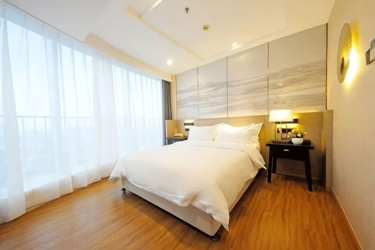 A hotel room with a large bed, a bedside table with a lamp, and a view of the outdoors through sheer curtains.