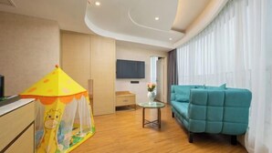 A hotel room with a wooden floor, a teal sofa, a small table with a vase, a TV mounted on the wall, and a play tent with a giraffe design. - Metropolo Jinjiang Hotel(Kunshan Jinying international Kunju theater store) (Kunshan)
